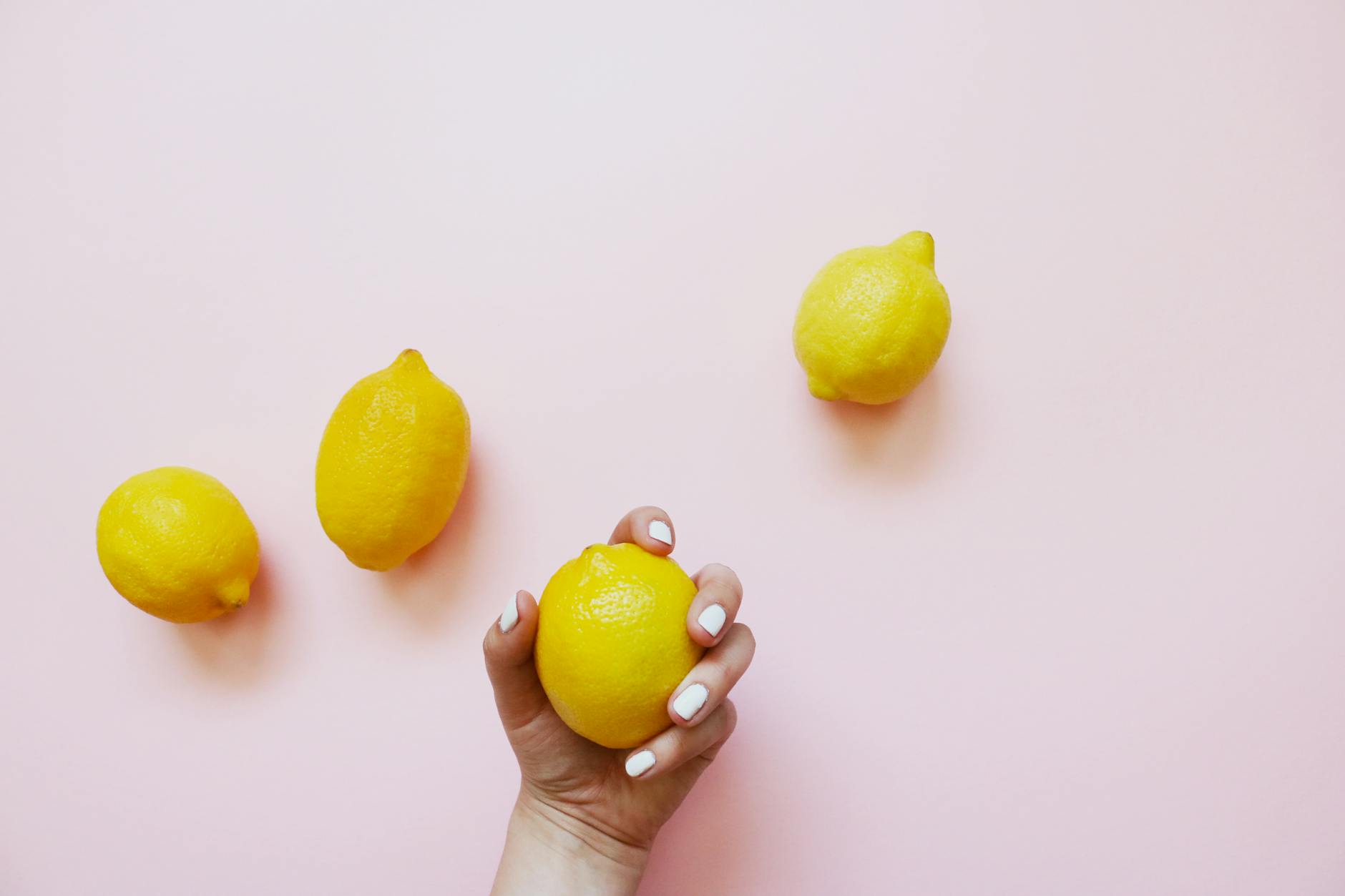 Eine Hand mit weißen Nägeln hält eine frische Zitrone vor zartem rosa Hintergrund.